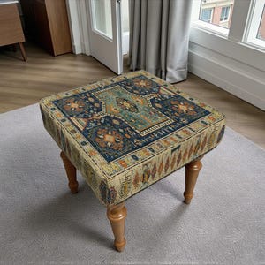 May include: Square ottoman with a patterned rug design in shades of blue, beige, and orange. The ottoman has four wooden legs and is placed on a grey rug. The rug design features floral and geometric patterns.