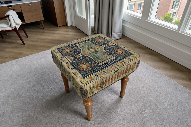 May include: Square ottoman with a patterned rug design in shades of blue, beige, and orange. The ottoman has four wooden legs and is placed on a grey rug. The rug design features floral and geometric patterns.