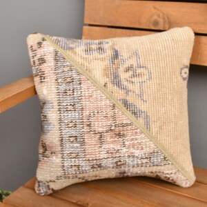 May include: Square decorative pillow with a beige and patterned design. The pillow features a diagonal seam, dividing the design into two sections. The pillow is placed on a wooden chair.