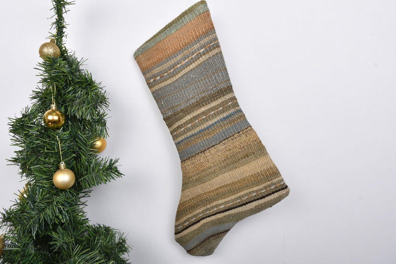 May include: A Christmas stocking with a striped pattern in shades of brown, tan, and blue. The stocking is hanging against a white background next to a small Christmas tree decorated with gold ornaments.