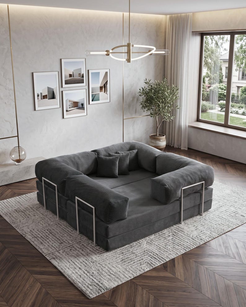 Puede incluir: Un sof&aacute; modular gris oscuro con un perfil bajo y reposabrazos redondeados. El sof&aacute; est&aacute; sobre una alfombra estampada y tiene soportes de metal plateado. Cojines decorativos y obras de arte enmarcadas completan el ambiente.