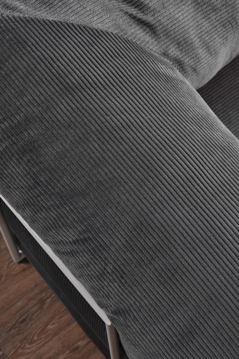 Puede incluir: Primer plano de una secci&oacute;n de un sof&aacute; de pana gris oscuro. La tela tiene una textura acanalada, con las crestas que corren diagonalmente a trav&eacute;s del marco. El sof&aacute; parece ser parte de un conjunto m&aacute;s grande, con un borde blanco y un marco de metal.