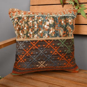 May include: A square cushion with a geometric pattern in shades of brown, orange, green, and blue. The cushion is placed on a wooden bench, showcasing its textured design and earthy tones. The cushion's dimensions are not visible.