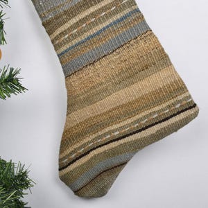May include: A woven Christmas stocking with a striped pattern in shades of tan, green, and blue. The stocking has a textured appearance and is decorated with white stitching. A Christmas tree with gold ornaments is in the background.