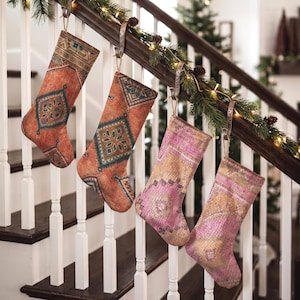 Könnte beinhalten: Vier Weihnachtsstrümpfe hängen an einem Treppengeländer aus Holz. Zwei Strümpfe haben orangefarbene und blaugrüne geometrische Muster, während die anderen beiden rosafarbene und goldene geometrische Designs aufweisen.