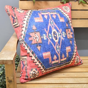 Könnte beinhalten: Quadratisches Zierkissen mit einem lebendigen, geometrischen Muster. Das Kissen hat ein blaues Mitteldesign mit orangefarbenen und weißen Akzenten, eingerahmt von einem roten Rand. Es liegt auf einer Holzbank.