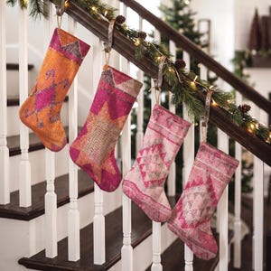 Può includere: Quattro calze natalizie decorative appese a una ringhiera di una scala in legno. Ogni calza presenta un motivo tessile intrecciato unico nei toni dell'arancione, magenta e rosa tenue, con disegni geometrici e bordi sfrangiati.