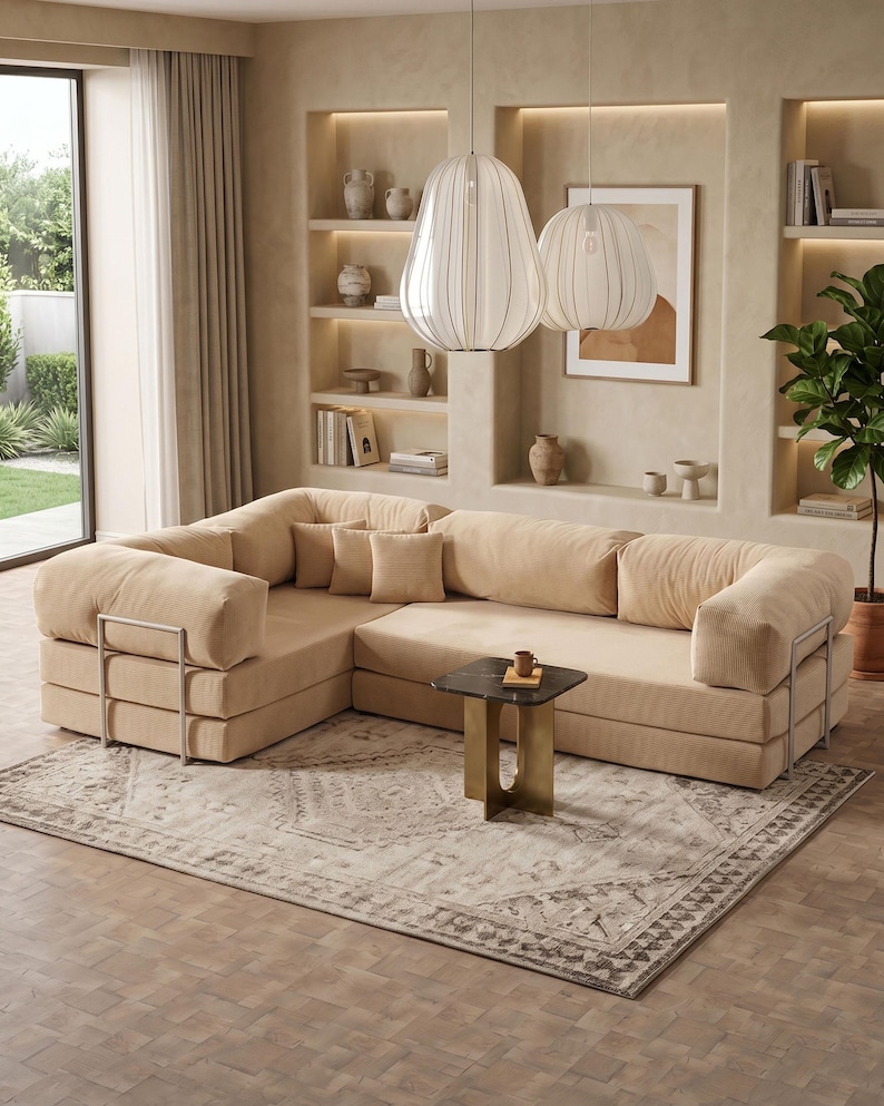 Puede incluir: Un sof&aacute; seccional beige con cojines cuadrados y detalles met&aacute;licos. El sof&aacute; est&aacute; sobre una alfombra estampada, con una peque&ntilde;a mesa auxiliar y dos l&aacute;mparas colgantes encima. La habitaci&oacute;n tiene estanter&iacute;as empotradas y una ventana grande.