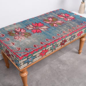 Könnte beinhalten: Eine Holzbank mit einem blauen, rosa und weißen gemusterten Kissen. Das Kissen hat ein traditionelles türkisches Kilim-Muster.
