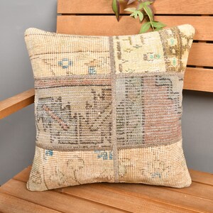 May include: Square decorative pillow with a patchwork design. The pillow features a mix of beige, brown, and blue tones, with a textured, vintage aesthetic. It is placed on a wooden chair, suggesting a home decor item.