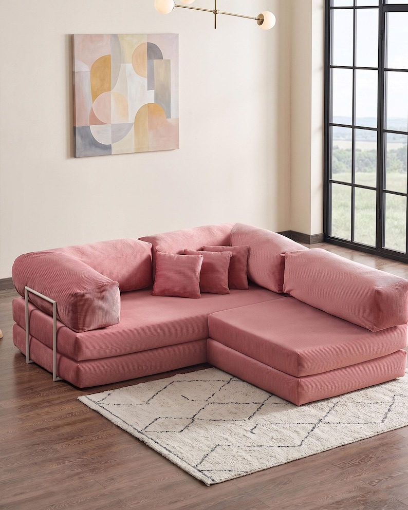 May include: A modular, sectional sofa in a dusty rose color. The sofa has a corduroy texture and includes several matching throw pillows. A metal frame supports the armrest. The sofa sits on a cream-colored rug with a geometric pattern.