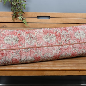 May include: A long, rectangular pillow with a red and cream floral pattern. The pillow is placed on a wooden bench. The pillow has a brown border and is likely used for decorative purposes.