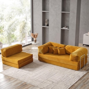 May include: A mustard yellow modular sofa and matching armchair set. The sofa has three square cushions and a metal frame. The set is placed on a light-colored rug with a geometric pattern. The room has a large window and neutral-colored walls.