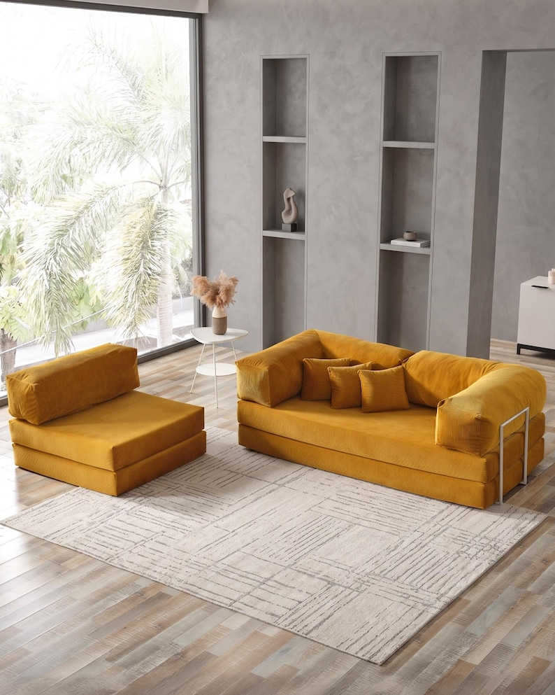 May include: A mustard yellow modular sofa and matching armchair set. The sofa has three square cushions and a metal frame. The set is placed on a light-colored rug with a geometric pattern. The room has a large window and neutral-colored walls.