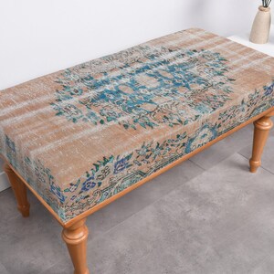 May include: A wooden bench with a beige and blue patterned cushion. The cushion has a floral design with faded colors. The bench has four legs and is made of wood.