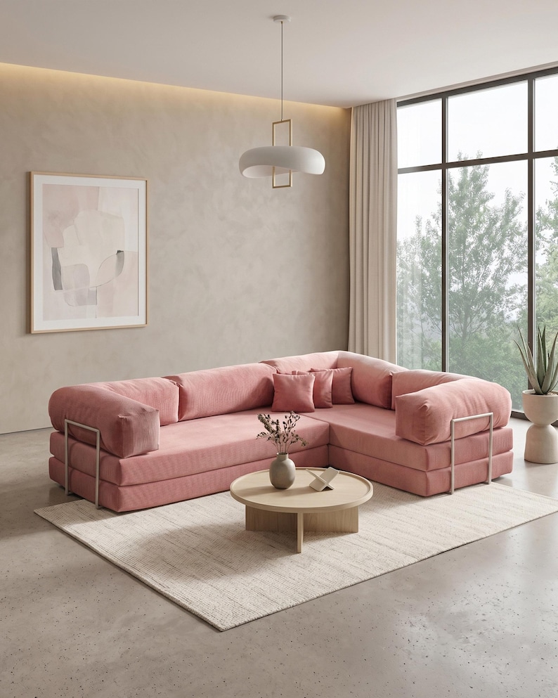 Puede incluir: Un gran sof&aacute; esquinero rosa con textura de pana y detalles met&aacute;licos. Una mesa de centro redonda de madera descansa sobre una alfombra crema. La habitaci&oacute;n cuenta con una obra de arte abstracta enmarcada y una moderna l&aacute;mpara colgante.