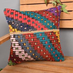 May include: A square, woven pillow with a vibrant, geometric pattern. The pillow features a mix of red, blue, teal, yellow, and purple hues. The pillow is placed on a wooden bench, showcasing its decorative design.