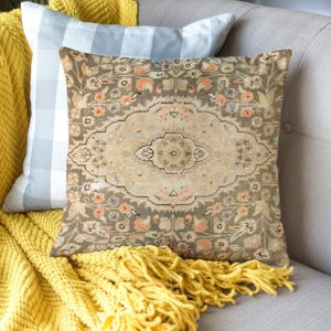 Könnte beinhalten: Ein dekoratives Kissen mit einem Vintage-Teppichmuster in Beige-, Grau- und Pfirsichtönen. Es liegt auf einem grauen Sofa, zusammen mit einer gelben Strickdecke und einem blau-weiß karierten Kissen.