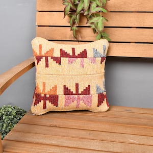 Pode incluir: Uma almofada quadrada decorativa com um padrão geométrico em tons de vermelho, laranja, rosa e azul sobre um fundo bege. A almofada é colocada num banco de madeira. O banco é feito de madeira castanha clara.