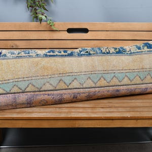 Pode incluir: Uma almofada longa e retangular com um visual vintage e desgastado. A almofada apresenta uma paleta de cores bege, azul e tons pastel, com padrões geométricos e um aspecto texturizado. Descansa sobre um banco de madeira, sugerindo um artigo de decoração para casa.