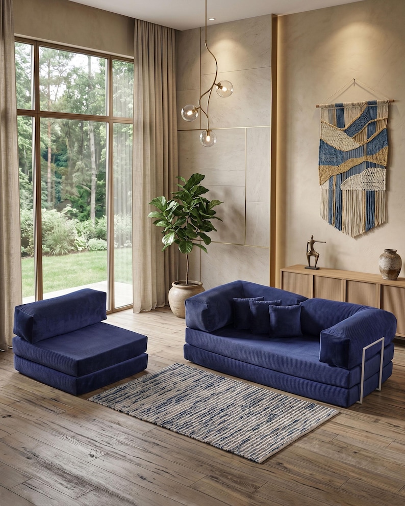 May include: A modern living room setup featuring a navy blue modular sofa and armchair. A patterned rug sits on the wooden floor. A large window with sheer curtains and a decorative wall hanging complete the scene. A potted plant adds a touch of nature.