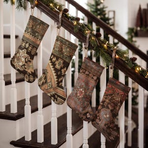 Könnte beinhalten: Vier Weihnachtsstrümpfe hängen an einem hölzernen Treppengeländer. Jeder Strumpf weist ein einzigartiges, kompliziertes Webmuster in gedeckten Erdtönen auf, darunter Braun-, Beige-, Salbeigrün- und verblasste Rottöne, die an Vintage-Teppichdesigns erinnern.