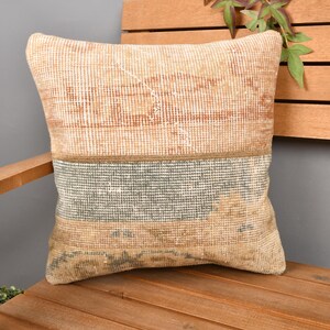 May include: A rectangular cushion with a patchwork design in shades of beige, brown, and grey. The cushion is placed on a wooden chair. The cushion's design features a striped pattern in the middle.