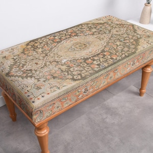 May include: A rectangular bench with a patterned top and wooden legs. The top features a detailed, vintage-style design in shades of green, beige, and orange. The bench has four turned wooden legs, adding to its classic aesthetic.