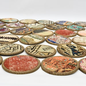 May include: A set of 20 round coasters made from colourful patterned fabric with a brown woven trim. The coasters are arranged in a semi-circle pattern on a white surface.