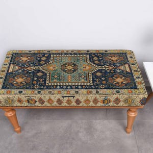 May include: A rectangular ottoman with a patterned top, featuring a central teal design surrounded by navy blue and gold accents. The ottoman rests on four light-coloured wooden legs. A small white side table with a vase of flowers is to the right.