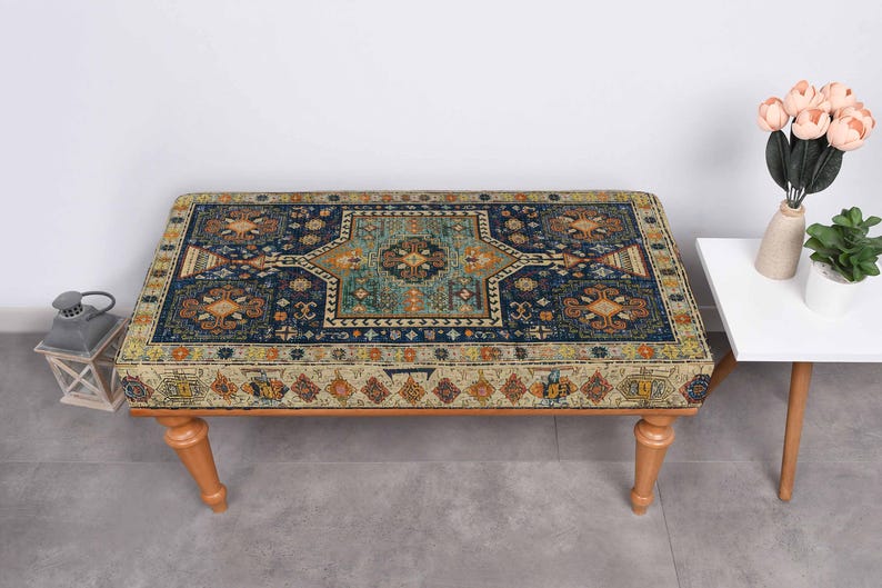 May include: A rectangular ottoman with a patterned top, featuring a central teal design surrounded by navy blue and gold accents. The ottoman rests on four light-coloured wooden legs. A small white side table with a vase of flowers is to the right.