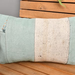 May include: Rectangular decorative pillow with a light blue and off-white color scheme. The pillow features a textured fabric with a central off-white panel flanked by light blue sections. The pillow is placed on a wooden surface.