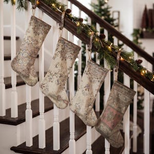 Pode incluir: Quatro meias de Natal penduradas num corrimão de escada de madeira. Cada meia apresenta um padrão de tapete vintage em tons suaves com motivos florais e geométricos complexos em bege, rosa seco, verde sálvia e azul desbotado.