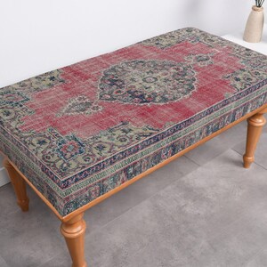Peut inclure: Un banc en bois avec un tapis rouge, bleu et vert à motifs sur le dessus. Le banc a quatre pieds et est en bois.
