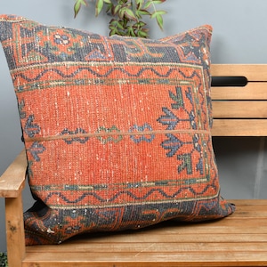 Op de afbeelding: Een vierkant decoratief kussen met een levendig, getextureerd, oranje en marineblauw patroon. Het kussen heeft een geometrisch ontwerp met bloemaccenten en ligt op een houten bank.