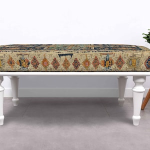 May include: A rectangular ottoman with a cream-coloured base and white, turned legs. The top features a patterned fabric with a beige background and geometric designs in shades of red, orange, blue, and yellow. The ottoman is a decorative furniture piece.