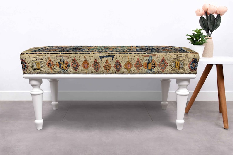 May include: A rectangular ottoman with a cream-coloured base and white, turned legs. The top features a patterned fabric with a beige background and geometric designs in shades of red, orange, blue, and yellow. The ottoman is a decorative furniture piece.
