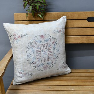 May include: A white throw pillow with a floral embroidered design in shades of pink, blue, and green. The pillow is sitting on a wooden bench.