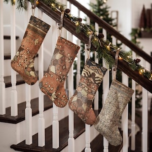 Pode incluir: Quatro meias de Natal penduradas num corrimão de escada de madeira. Cada meia apresenta um padrão tecido único e complexo em tons de ferrugem, castanho, bege e verde suave, lembrando designs de tapetes antigos com motivos geométricos.