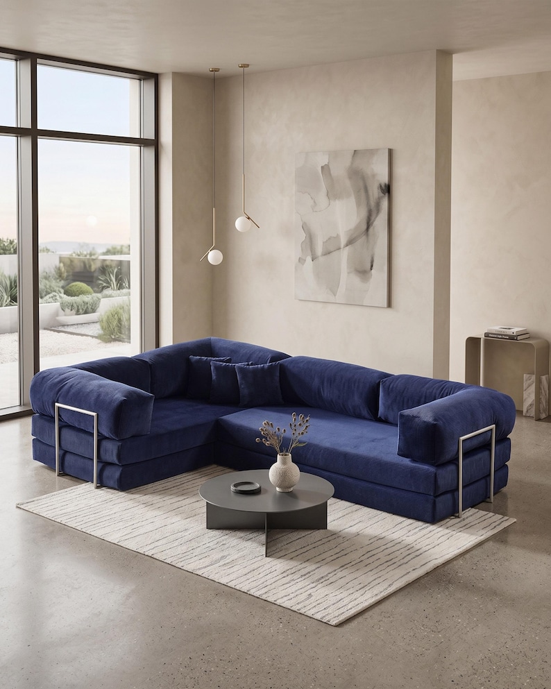 Puede incluir: Un moderno sof&aacute; seccional azul marino con detalles met&aacute;licos plateados. El sof&aacute; est&aacute; sobre una alfombra clara, con una mesa de centro gris oscuro y un jarr&oacute;n con flores secas. La habitaci&oacute;n tiene grandes ventanales y paredes de tonos neutros.
