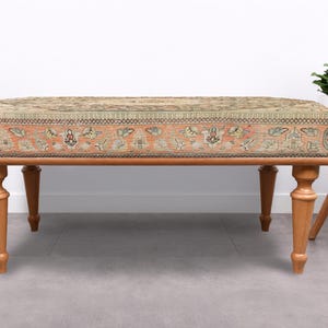 May include: An upholstered bench with a patterned top in shades of beige, orange, and green. The bench has four turned wooden legs. A small white side table with a vase of flowers is to the right.