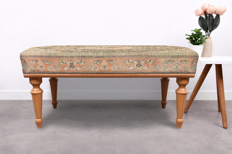 May include: An upholstered bench with a patterned top in shades of beige, orange, and green. The bench has four turned wooden legs. A small white side table with a vase of flowers is to the right.
