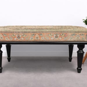 May include: An upholstered bench with a rectangular top featuring a patterned design in shades of beige, orange, and green. The bench has a black wooden frame and four turned legs. A small white side table and decorative flowers are in the background.