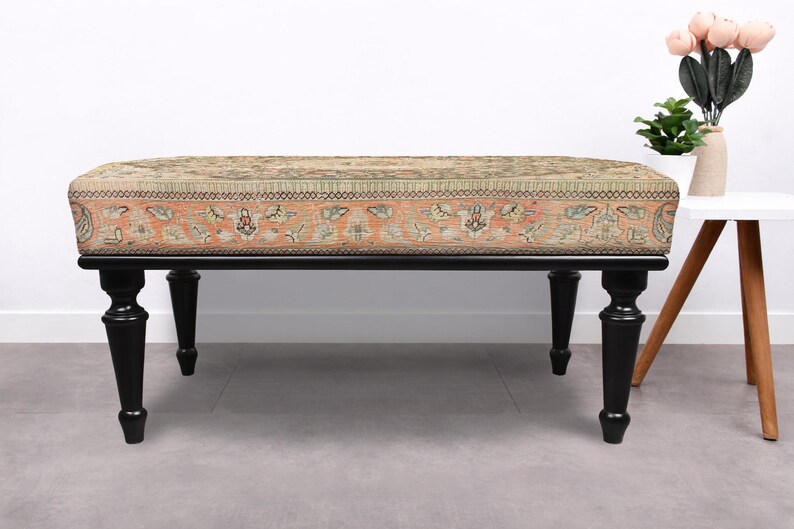 May include: An upholstered bench with a rectangular top featuring a patterned design in shades of beige, orange, and green. The bench has a black wooden frame and four turned legs. A small white side table and decorative flowers are in the background.