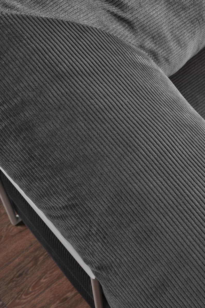 May include: Close-up of a section of a dark gray corduroy sofa. The fabric has a ribbed texture, with the ridges running diagonally across the frame. The sofa appears to be part of a larger set, with a white trim and metal frame visible.
