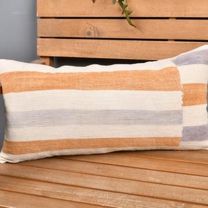 May include: A rectangular cushion with horizontal stripes in orange, cream, and grey. The cushion is made of woven fabric and rests on a wooden bench. The design adds colour and texture to the setting.