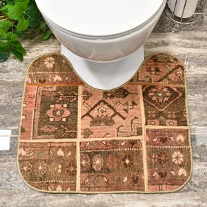 May include: A U-shaped bathroom rug featuring a patchwork design of various brown and dusty pink geometric patterns. The rug has a finished edge and is positioned around the base of a white toilet on a gray stone-look tile floor.