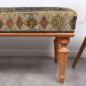 May include: A rectangular bench with a patterned top and wooden legs. The bench top features a beige background with a repeating pattern of diamonds and other shapes in shades of orange, blue, and yellow. The legs are light brown.