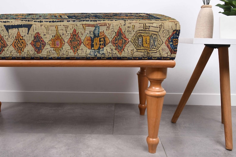 May include: A rectangular bench with a patterned top and wooden legs. The bench top features a beige background with a repeating pattern of diamonds and other shapes in shades of orange, blue, and yellow. The legs are light brown.