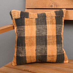 May include: Square decorative pillow with a black and orange plaid pattern. The pillow is made of a woven fabric and is placed on a wooden surface. The pillow's design features a classic checkered pattern.
